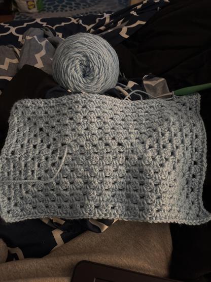 A light blue crocheted piece is laid out on a bed, accompanied by a coordinating ball of yarn and a crochet hook. Various fabric patterns and colors can be seen in the background, suggesting a cozy crafting environment.