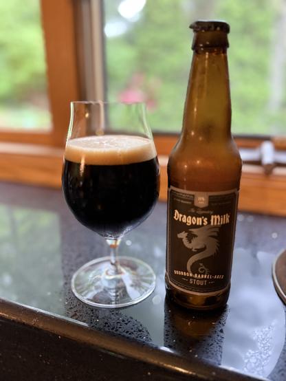 A tall snifter glass of dark beer with a dark tan head. The frosty brown bottle is to the right with a black label, white lettering, and a silver dragon in silhouette