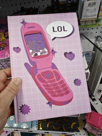 Photo of a notebook with a pink flip phone like from the 2000s. Also the phone says "lol"