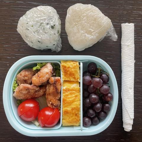 Child’s Japanese packed lunch - plain rice ball, seasoned rice ball, chopsticks wrapped in paper, and a pale blue lunch box containing fried chicken (karaage), lettuce, cherry tomatoes, sweet omelette (tamagoyaki), and a small bunch of tiny purple grapes (Japanese ones where the skin pops off)