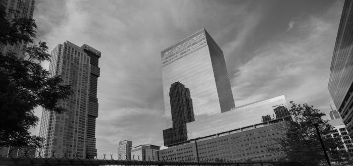 A monochrome wide-angle photo of some urban buildings: a little tilty, with the tops of the buildings trying to converge toward the top.  The sky is dramatic, and the top of a chain-link fence runs along the bottom of the photo.  One of the buildings is particularly reflective, and on it we see the image of another nearby tower.