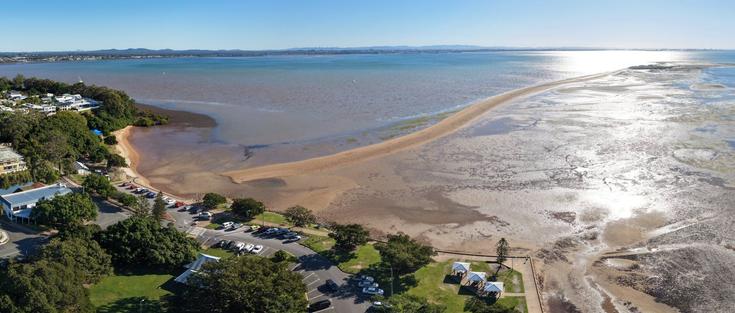 A large sand spit extending out in to the water from parkland. It's a bright, sunny day, the light of the sun is reflecting off of the water
