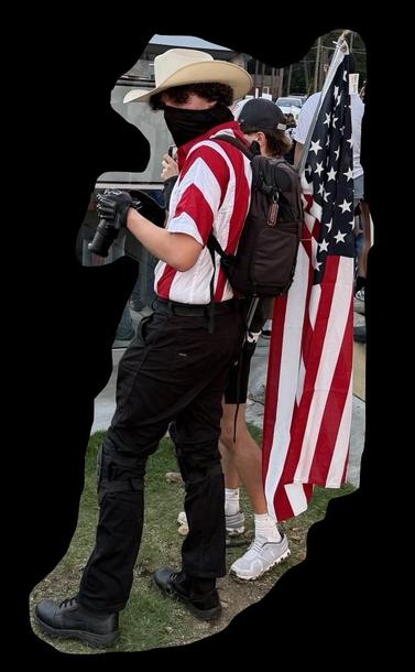 man in white cowboy hat, red and white striped shirt and carrying american flag. Black mask.