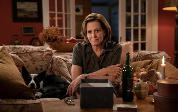 Sigourney Weaver sits in a couch alone in a living room. A bottle of wine is on the table in front of her.