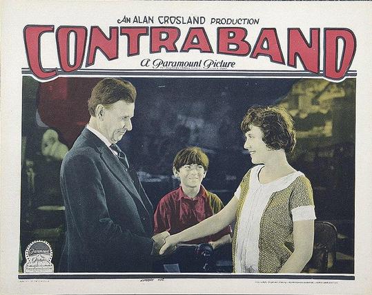 A poster of the film. A man and a woman cross hands while a boy in the middle of them sees it. The man wears a black suit. The woman wears a short-sleeved white and yellow dress. Her hair is short and dark. The boy is wearing a long-sleeved, red shirt. His hair is short, straight and dark.