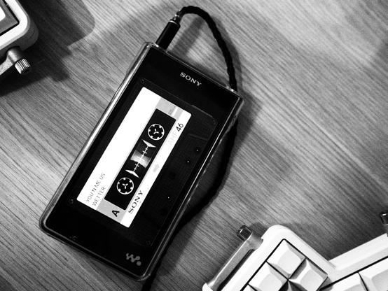 Black and white photo of a sony walkman. The screen is showing an animation of a cassette tape being played. The name of the artist and song is displayed (Wetter - You N Me Us).
