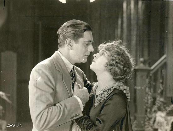 A still of the film. A man and a woman are about to kiss. His hair is short, straight and dark. He is wearing a gray suit, a stripped tie and a stripped shirt. She has wavy, short, brown hair. She is wearing a long-sleeved black dress.