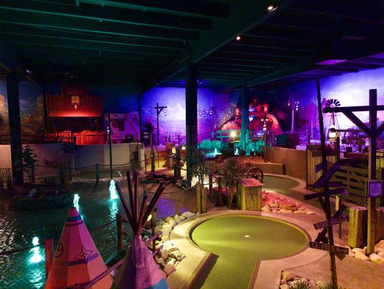 Indoor miniature golf course with dramatic lighting featuring a large circular putting green in the foreground surrounded by artificial rocks. A colorful teepee structure sits to the left near a water feature with illuminated fountains. The space has exposed beams on the ceiling, vibrant purple and blue lighting, and themed decorations including windmill structures and rustic wooden elements throughout the multi-level course.
