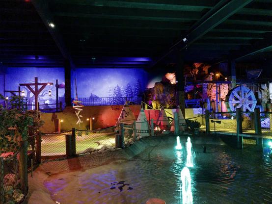Multi-level indoor miniature golf course viewed from an elevated walkway with fencing. The scene shows various themed holes with Western and frontier motifs, including wooden structures, painted backdrops depicting landscapes, and a large water wheel. Dramatic colored lighting in blues, purples, and greens creates an atmospheric environment with water features containing illuminated fountains below.