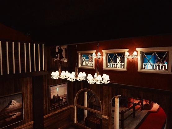 Elegant wooden interior of an upper level viewing area with rich dark wood paneling and ornate chandeliers with lampshades. Large windows with decorative crosshatch patterns look down onto the miniature golf course below. The space features a stone fireplace and western-themed artwork on the walls, creating a lodge-like atmosphere with warm ambient lighting.