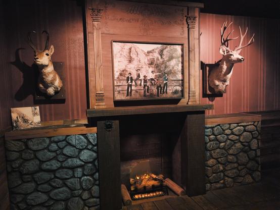 Rustic fireplace display with stone base and wooden mantel featuring mounted taxidermy including deer antlers and a pronghorn. Above the mantel hangs a framed painting depicting a western scene with cowboys or frontier figures. The fireplace contains artificial logs with orange lighting to simulate flames, and the overall aesthetic emphasizes a hunting lodge or Old West theme.