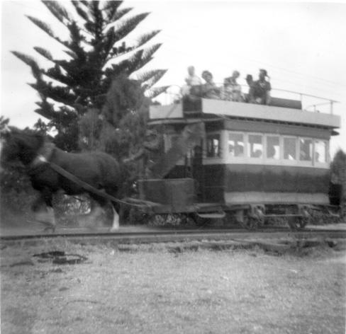 A black and white photo of a horse tram.  A large horse at left is hauling a double deck 4-wheeled tram which has an enclosed saloon on the lower level connected to an open topped upper lever by a curved staircase at the front of the tram.  The top deck has a fairly flimsy looking handrail running around the edge and a knifeboard longitudinal seat in the centre.    The underframe of the tram appears to have a significant bend in it meaning the driving position at front is sagging towards the roadway.