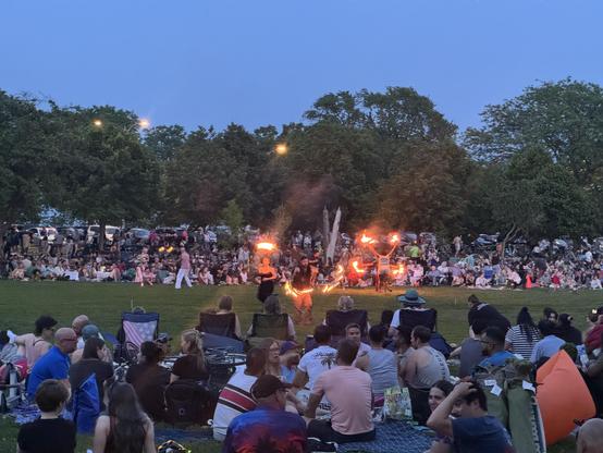 A lively outdoor gathering in a park during dusk, featuring a crowd watching fire performers. Several people are sitting on blankets and lawn chairs, while performers hold flaming torches. The scene has a festive atmosphere with greenery and cars in the background.
