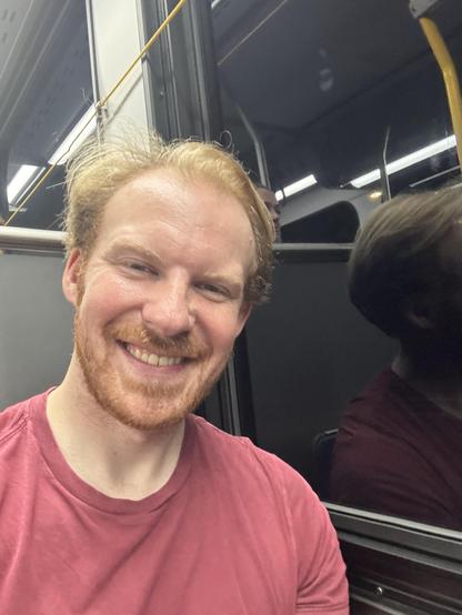 A smiling person with light red hair and a beard is sitting in a bus, wearing a maroon shirt. The bus interior is visible in the background, including a reflection in the window.