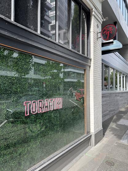 Outside windows at Tora Tiki. It is completwlyncoveree in fake vegetationnon rhebimaidenwirh a neon “ToraTiki” sign.