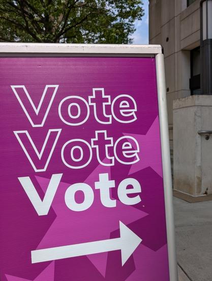 A purple sign next to one of Arlington's early-voting sides reads "Vote Vote Vote," with the county's Courthouse office building in the background.