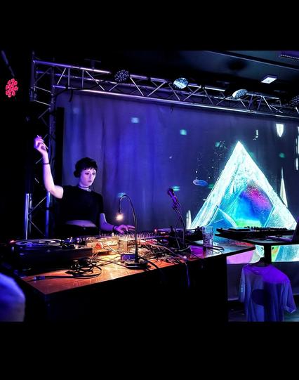 A serioused faced girl with her hands up controlling dj equipment next to a psychedelic crystal image