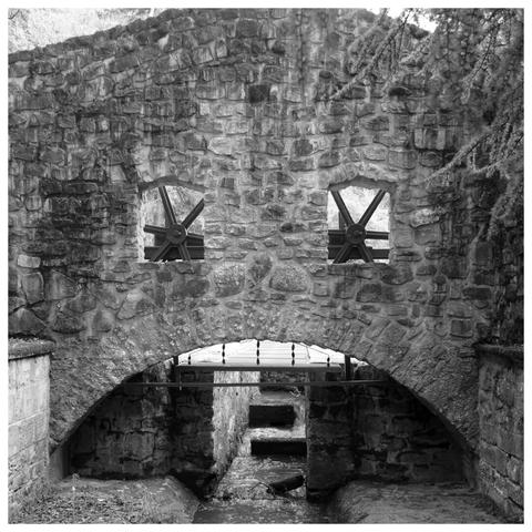 Black and white photo taken in the Alzette Valley. A bridge over the river with two wooden mills look like the face of a monster. The arch above the water is the unhappy mouth of this old monster made of stones.