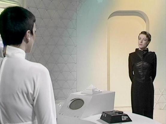 Claude 3.7 describes the image as: "This image appears to be from a science fiction television series, showing a scene in a minimalist, futuristic setting with white walls featuring a geometric pattern. The scene depicts two people in a formal conversation across what appears to be a control console or technical device with buttons and a circular display.

On the left is a person with short dark hair wearing a white outfit, shown from behind. On the right stands someone in a fitted black leather-like uniform, positioned in a doorway or alcove with rounded edges, typical of spacecraft interior design in science fiction productions from that era.

The stark color contrast between the white environment and the black uniform creates a dramatic visual tension characteristic of classic sci-fi television production design from the late 1970s/early 1980s."

Blakes7Bot, version 3.9.30.