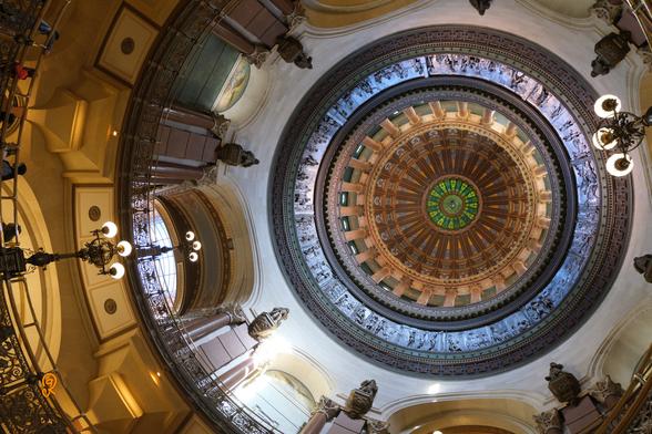 Looking up at the dome of the Springfield Illinois USA capitol building. Part of the railing of the middle and upper floors are visible around and to the left. The dome goes from green, brown, golden, sky blue, alabaster, to golden colors as it descends from topmost to the visible bottom rings.