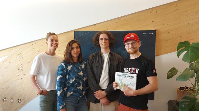Indoor group photo of for people standing in front of a wall. Three of them are worker-members of the workers cooperative Analyse og Tall, the fourth one is Jacob (me). I'm wearing a Red party cap which is covered in pen markings left by the cool people I've met along the way. I'm holding a book called "kooperativ håndbog" in front of me, which Konrad of Analyse og Tall kindly lends me