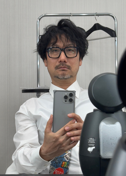 mirror selfie of Hideo Kojima, his neck is pushed back a little bit and he's wearing a white suit with a tie that most of his arm is covering, while staring at himself in the mirror.