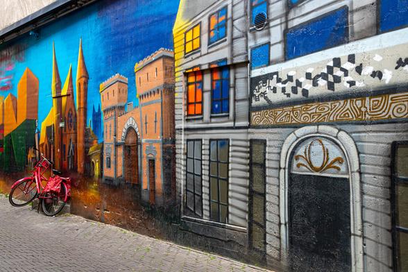 A colorful mural. The wall goes from right to left. At the beginning a gray house with doors and windows, then an orange castle and at the end a red fantasy castle with high red pointed towers. Above it is a blue sky. Against the red tower at the end stands a red painted bicycle, a real one. Bottom left in a slanted point of the street.