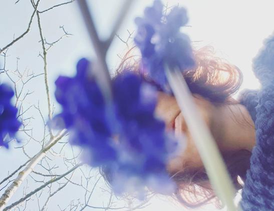 Femme penchée au dessus d'une fleur bleutée. Son visage est à demi caché entre les pétales et son pull de la même couleur