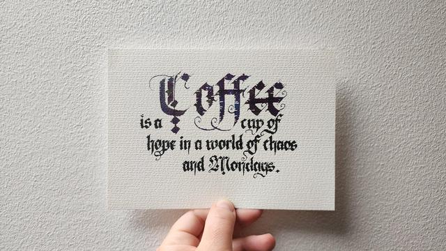 Calligraphed in fancy Blackletter script on textured white paper;

'Coffee is a cup of hope in a world of chaos and Mondays'

The word 'Coffee' is big and ornate and in sparkly blue-purple ink, the rest is in smaller lettering in black ink.