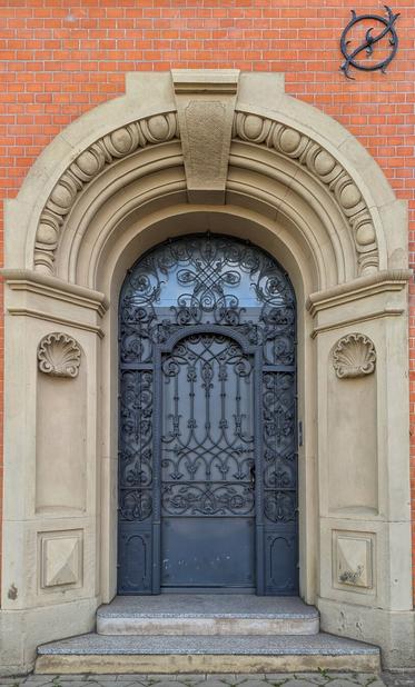 An ornate, arched, dark grey wrought-iron door is set within a lighter grey stone archway, framed by brickwork. The stone archway is intricately carved with a shell motif on either side of the base, and a textured, rounded pattern along the arch. The top of the archway features a decorative rectangular keystone. The wrought-iron door itself has elaborate scrolling patterns and a glass pane at the top. The building's red brick wall is visible around the archway, and a dark, abstract metal sculpture is affixed to the brickwork in the top right corner. Three stone steps lead up to the door.