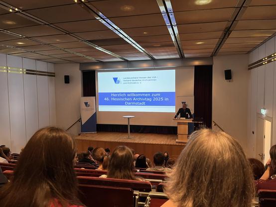 Vor einem Plenum in aufsteigenden Sitzreihen wird eine Folie mit der Aufschrift „Herzlich Willkommen zum 46. Hessischen Archivtag 2025 in Darmstadt“ gezeigt.