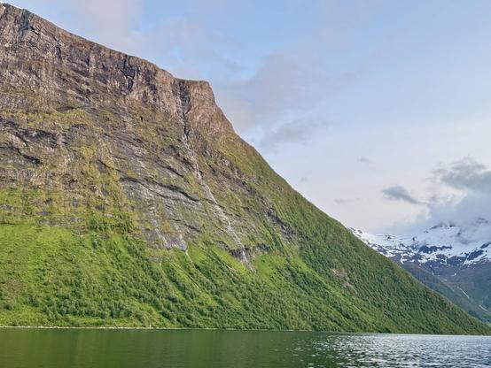 A mountain angles down from top left to bottom right of the picture frame. The top half is a brownish grey color of a slate rock face, the bottom half is a rich green from the trees. It meets a fjord, which reflects the green of the trees. The sky is a pinkish blue of the golden hour sun, which lasts from about 11 pm til 3 am rather than an hour.