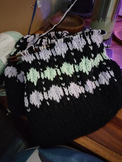 A knitting project to create a shirt on a wooden desk in the corner of a room. There is a diamond -pattern in light purple and light mint alternating between rows, with black in-between. All through only two are finished and the 3rd is close to being finished. 
The needles are dark wood with cable in-between. 
There are a little bit of clutter, a waterbottle with scraped off green paint, coasters, a brown candle, a brown folder leaning to the wall.