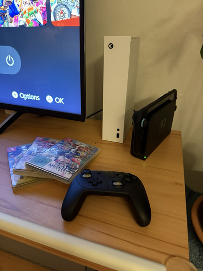 Foto von einer Xbox neben einer Nintendo Switch 2 auf einem TV Sideboard. Daneben drei Nintendo Switch Spiele, ganz oben Super Smash Bros