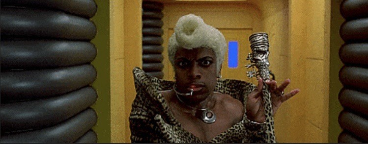 Radio personality Ruby Rhod of The Fifth Element shortly before he responds to the question "Green?" with "Supergreen!" whilst being held by the neck by a displeased live on-air guest.

Ruby stares intently towards the camera after just arriving on-scene. He has white hair upswept into a cylindrical bun just above his forehead, a lowcut, skintight animal-print suit with a tall collar that is almost off-the-shoulder, wearing a silver pendant on a choker and a headset with a red light nwar his mouth indicating his radio feed is live. Between his left index finger and thumb he delicately holds a sceptre with an ornate tip. The pattern on the body of the sceptre matches Ruby's suit.