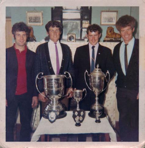Slightly faded scan of a square format colour photograph taken at some point in the early 1970s most likely. It's the interior of a house with four men showing off won trophies in front of them. The rear wall is painted pale blue and there's a tall white mantelpiece behind the men here with a small clock and small statues on its top as well as a larger mirrored unit in the centre and two framed photos hanging on the wall either side. The four men are standing side by side, all with similar looks. They're all white, smiling, with dark, curly hair - the man on the right has a larger mass of it - and all are wearing dark trousers, with two sporting dark cardigans over light shirts and ties, and the other two have jackets, with one having a light shirt and tie and the other wearing an open-necked, red, collared top. A small, low table is set in front of them with a white cloth on it, and on that are three large silver trophies and a couple of smaller trophies and shields resting in front of them.