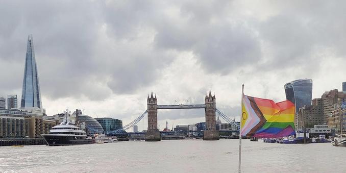 Picture of tower bridge taken from a ferry. The ferry's rainbow flag is showing in the foreground.