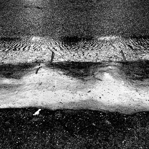 A close-up of a wet road surface, showing ripples in the water and a contrasting textured curb in black and white.