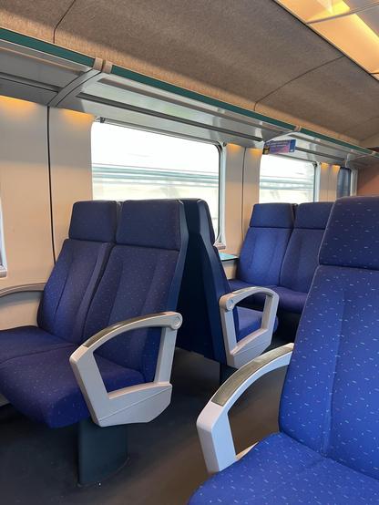 Innenraum eines Zuges mit blauen Sitzgelegenheiten mit Armlehnen und großen Fenstern. Die Umgebung ist hell und zeigt moderne Designelemente.