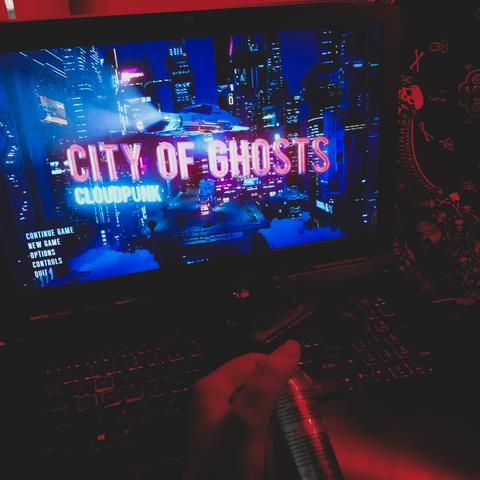 cloudpunk expansion city of ghosts loading screen or main menu on a laptop, all lit with red light. there's an xbox controller atop the keyboard, and a hand nearby holding a canned drink.