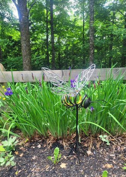 Une abeille en acier revalorisé, de grande dimension est fixée sur une barre de 24" de haut. L'abeille est devant un bosquet d'iris, devant une clôture en bois. En arrière-plan, il y a la forêt.