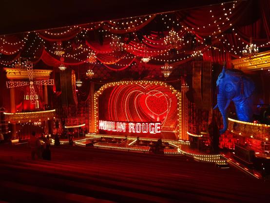 Overview of the Moulin Rouge musical stage. The main stage is decorated by concentric stage-wide red hearts. On both sides of the stage, there is a balcony and additional decorate (a windmill left and an elephant right). In front of the stage, a catwalk extends lengthwise towards the audience, at the end of it it splits in two catwalks towards both sides of the stage. Above these catwalks text "Moulin Rouge" is suspended in illuminated letters. The ceiling is decorated with hanging lights. Between the catwalks, small tables and chairs are placed for audience to watch.

The room is mostly empty, a few people are walking around.

Overall coloring is red and gold, with lots of lights and frilly details.