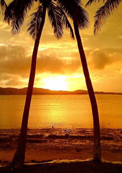Photo shows a sunset between two palm trees crossing each other at the top, framing the setting sun between the trunks. The main colour is yellow, orange and bronze. At the bottom of the photo the palm trees are planted in earth on the border of a beach. The beach stretches into the sea, with stretches on with only small ripples of disturbance in the surface from the afternoon breeze. In the background another island harbouring the water body with small hills stretching from the near area to the left, forming the horizon where the sun sets. The sky is lightly clouded to the right, but a thicker cloud system is coming from the left warning of a possible rain shower during the night.