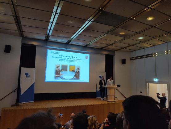 Vor einem Plenum auf einer aufsteigenden Sitzreihe stehen zwei Personen an einem Pult auf einem Podium. Ein Präsentationsfolie zeigt die Aufschrift "Ganz neue Töne. Ein Werkstattbericht zum Podcast Projekt des Hessischen Landesarchivs".