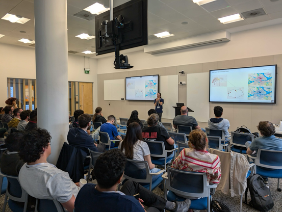 Crowd shot from the rear of the room, showing Fernando speaking