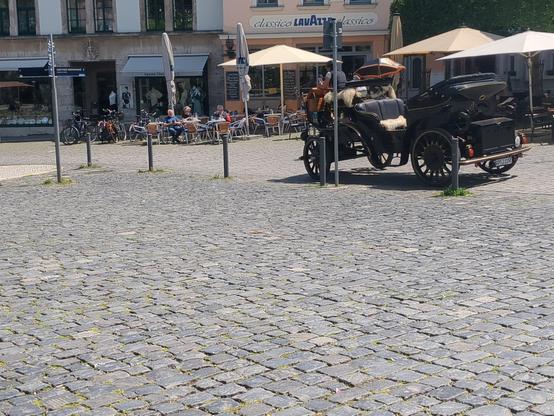 Photo of an cobble stone place in a city with old buildings around. On the place stands a horse cart without horses.