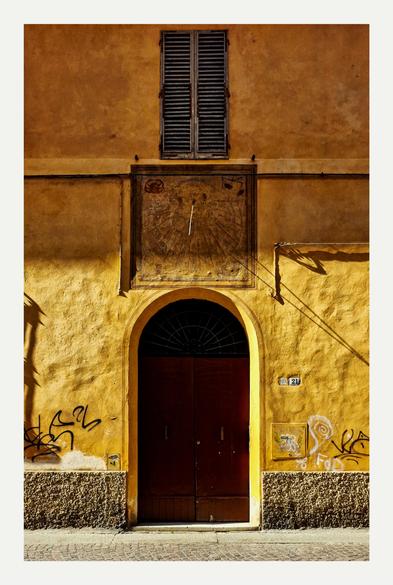 The photo is a section of a sunny  ochre/yellow building. We are focused on a weathered, arched doorway with a dark brown door. Above the door is a square sundial, painted directly onto the wall. On the sundial you can see a crab on the top left of the sundial and some scales on the top right. There is a closed wooden window with shutters above the sundial. The wall shows signs of age and some graffiti near the bottom.