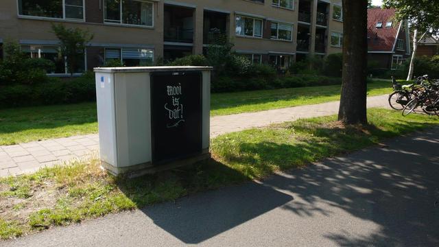 elektriciteits kastje voor flat, voor het voetpad en langs een weg met boom rechts ervan en geparkeerde fietsen.
Reclamebord zwart gemaakt en met sierlijke witte letters: Mooi is dat! er op. Het bord staat in zijn eigen shaduw en die zie je ervoor op de grond