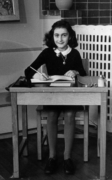 Anne Frank seated at a desk with an open book and a pen, looking directly at the camera, with a bottle of ink by her side.