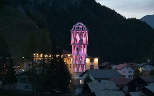Night time photo of 3D printed tower at the center of a small Swiss mountain village. The tower is made from pale concrete, illuminated in pink LED lights. Steep mountain slopes rising up behind the village